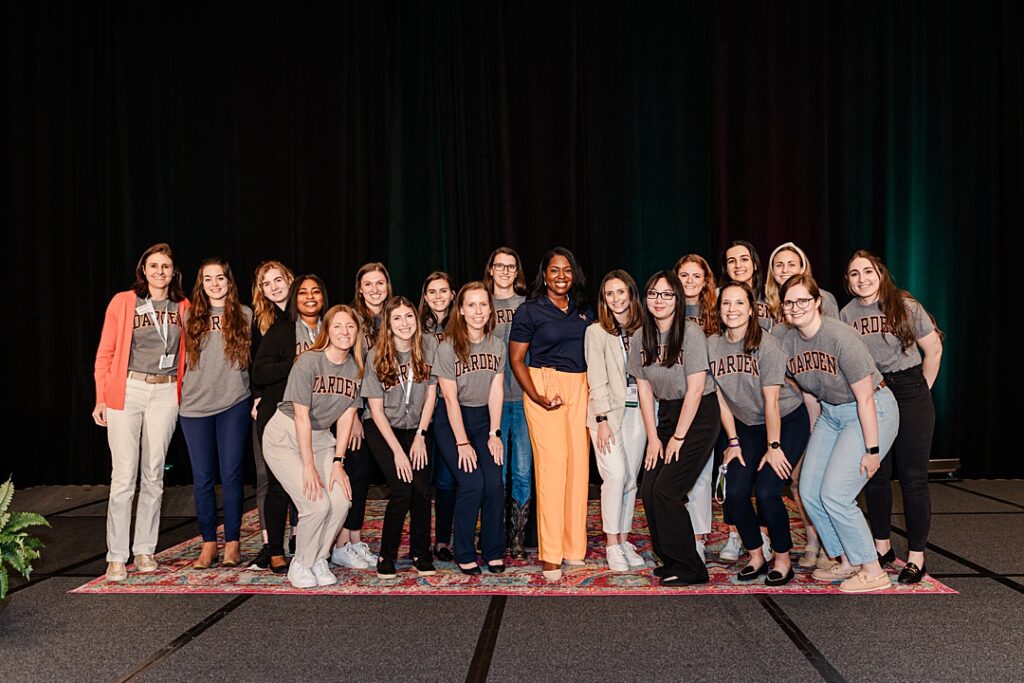 A group of women smiling and posing for a group shot during the Forte Foundation's MBA Women's Leadership Conference in Dallas, Texas. Most of the women are wearing grey shirts with a large logo of their university while the woman in the middle is wearing a navy polo with a small logo of their university.