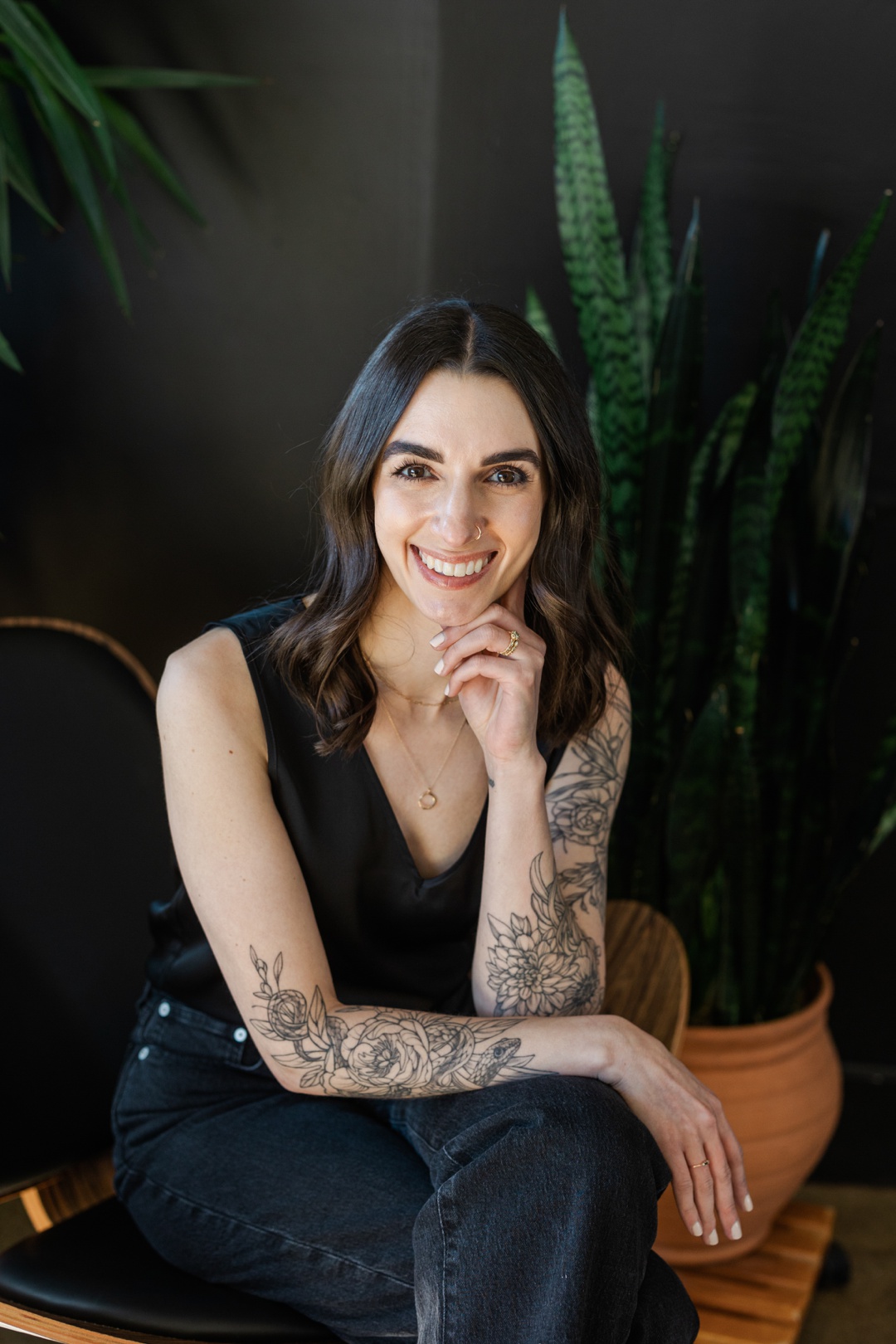 A smiling woman resting her hand on her chin with crossed legs in front of a black background and plants
