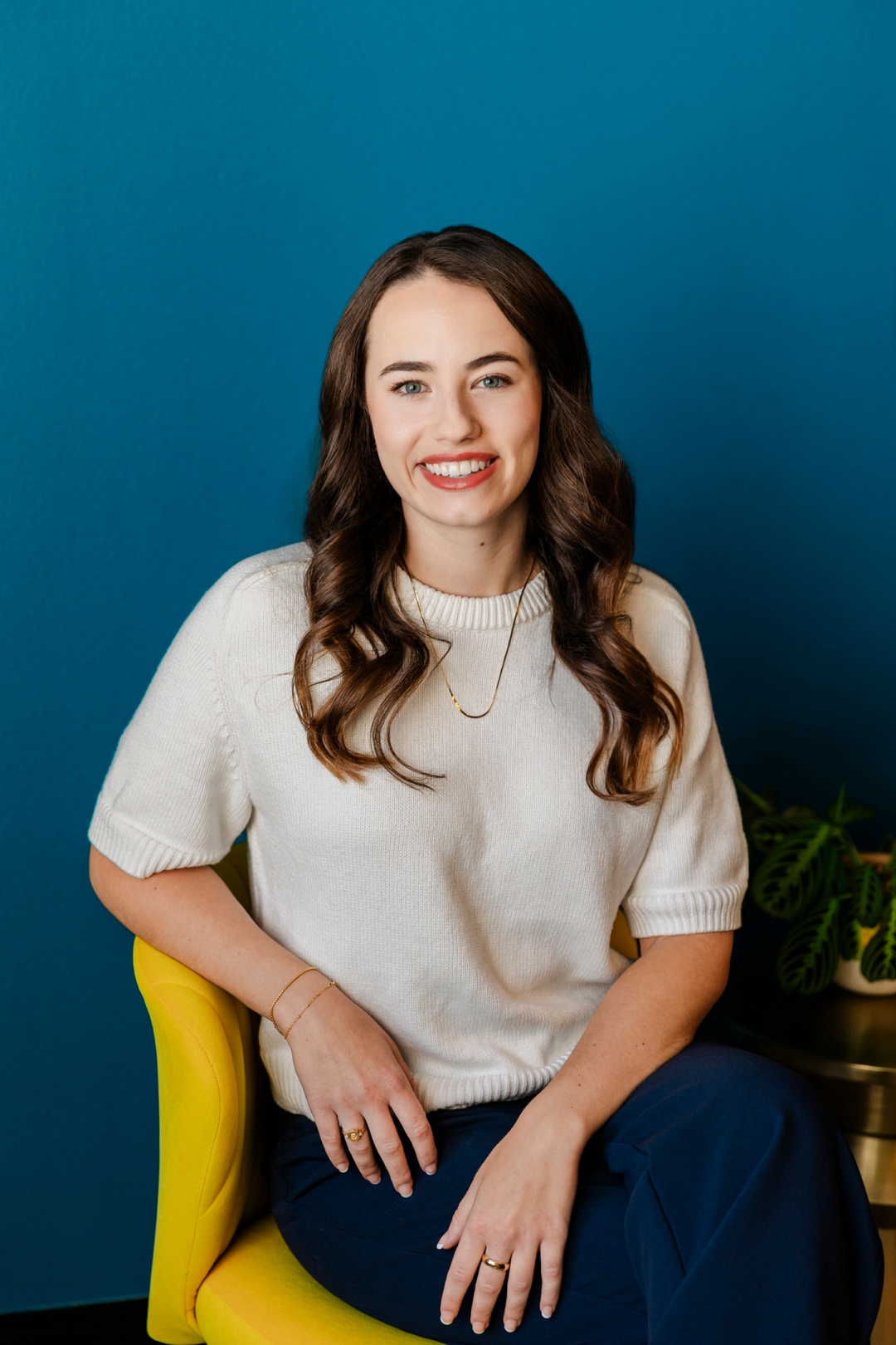 A smiling woman sitting on a yellow chair in front of a blue background