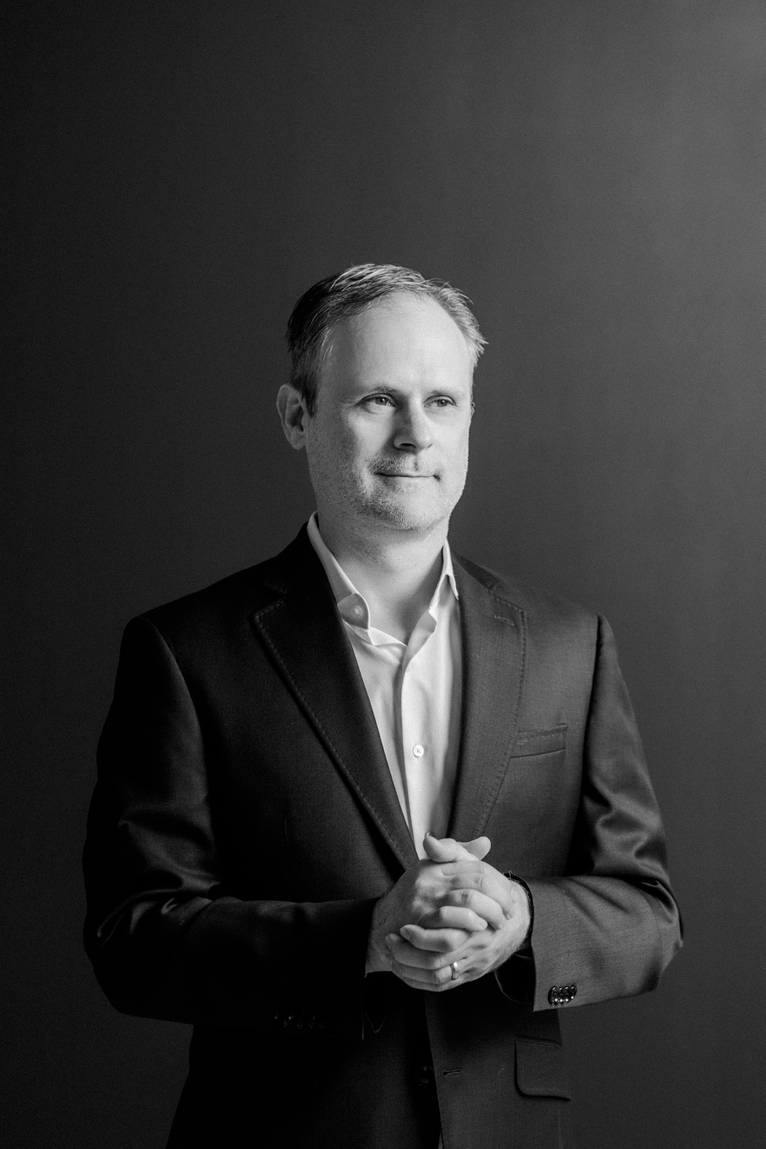 A black and white photo of a man clasping his hands and standing in front of a dark background