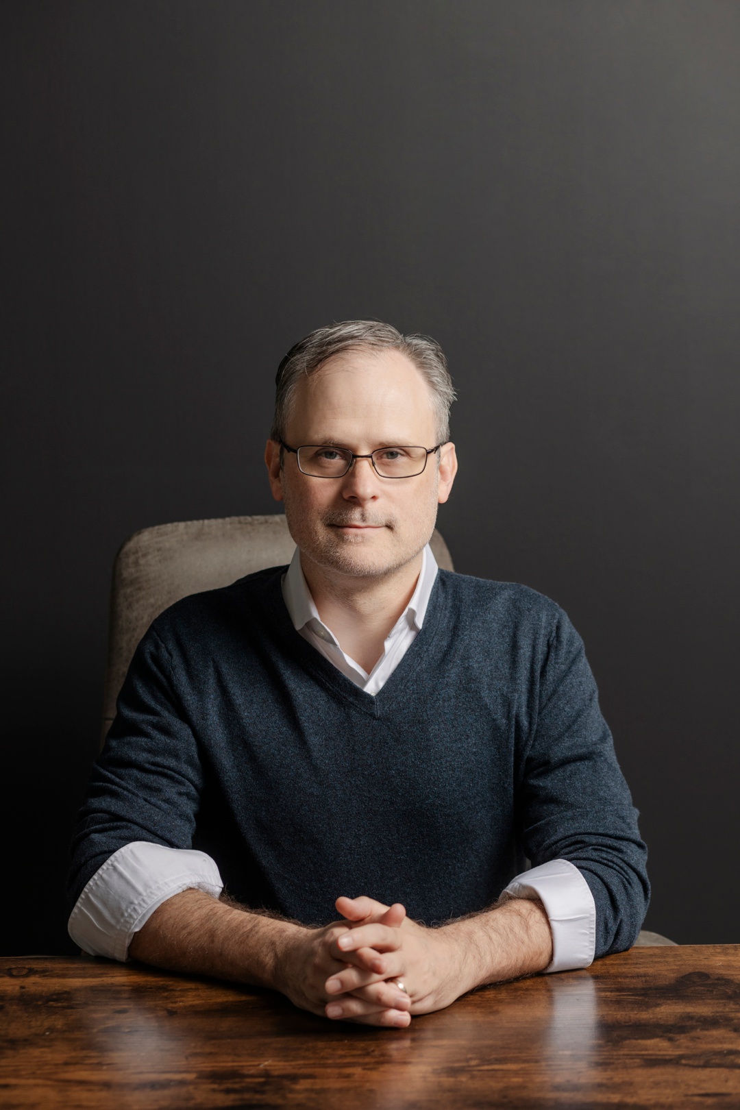 A man subtly smiling sitting in a chair with clasped hands resting on a desk