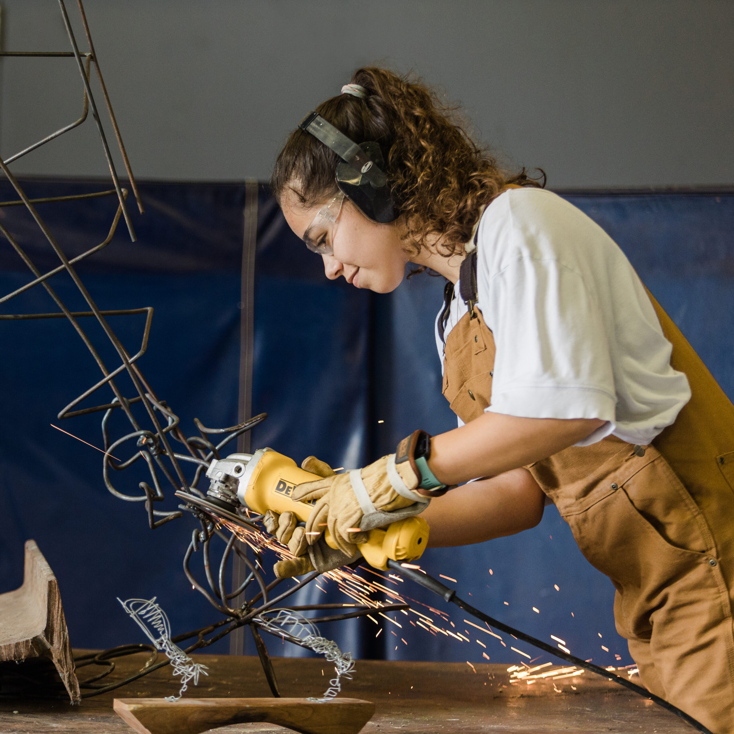 DFW brand photographer; photo of a woman wearing brown denim overalls, work gloves, and safety goggles using a power grinder on a metal sculpture in front of a dark blue background