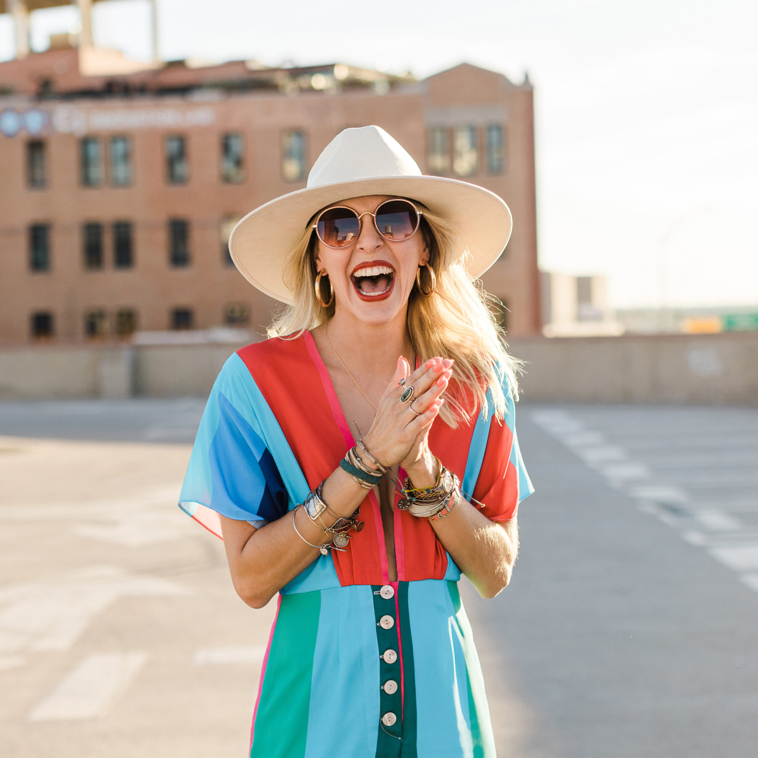 DFW Branding Photographer: a woman laughing and hands together on top of a parking garage. She's wearing a cowboy hat with large sunglasses, a colorful dress, and lots of jewelry.