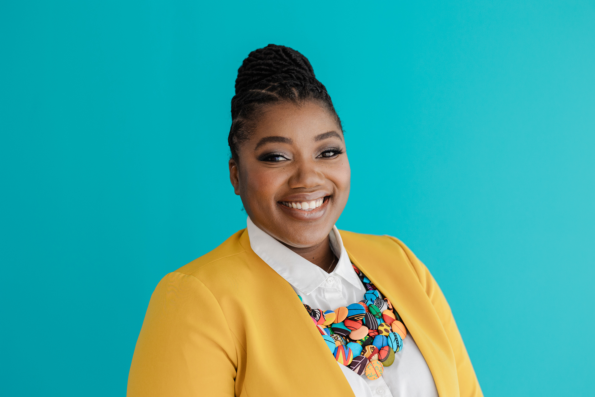 DFW Branding Photographer: a woman smiling and posing in front of a blue background. She's wearing a white dress shirt, yellow blazer, and a necklace made of colorful buttons.