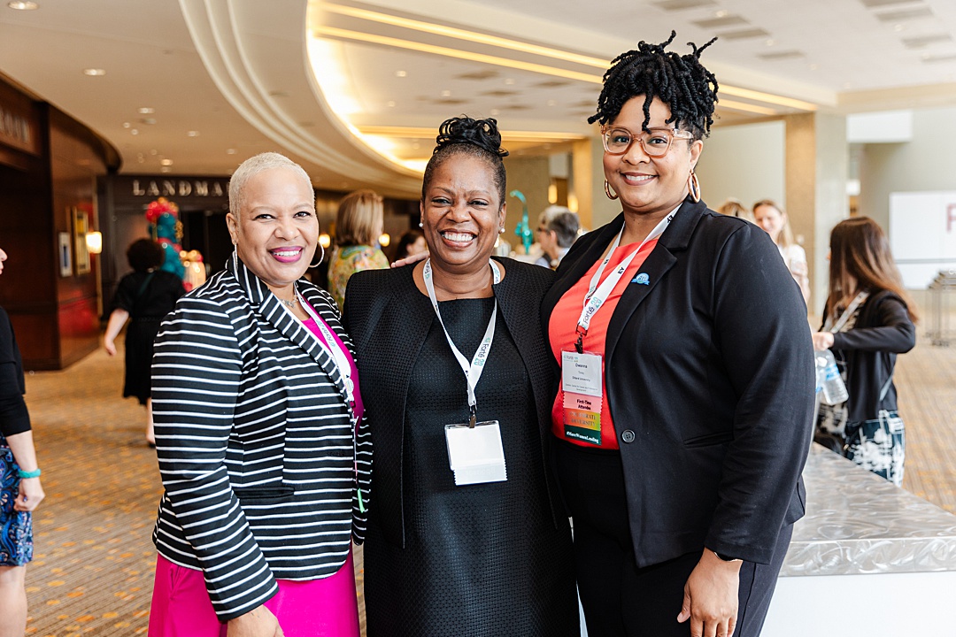 DFW Event Photographer: three women smiling and posing together during the Forte Foundation's MBA Women's Leadership Conference in Dallas, Texas. All three women are dressed in varying types of professional business attire and have on Forte branded lanyards with name tags.
