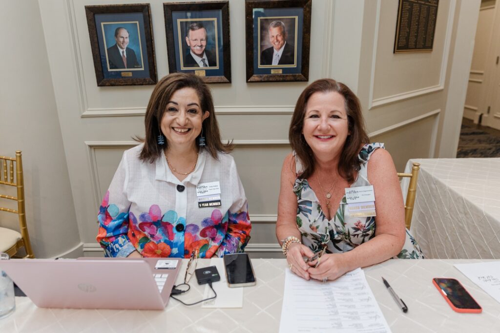 Two caucasian women members of the Society of Wedding Professionals wearing floral tops, smiling, sitting, and working at a reception desk.