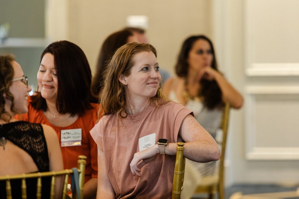 A candid shot of a member of the Society of Wedding Professionals watching a presentation while wearing a salmon top and smiling.