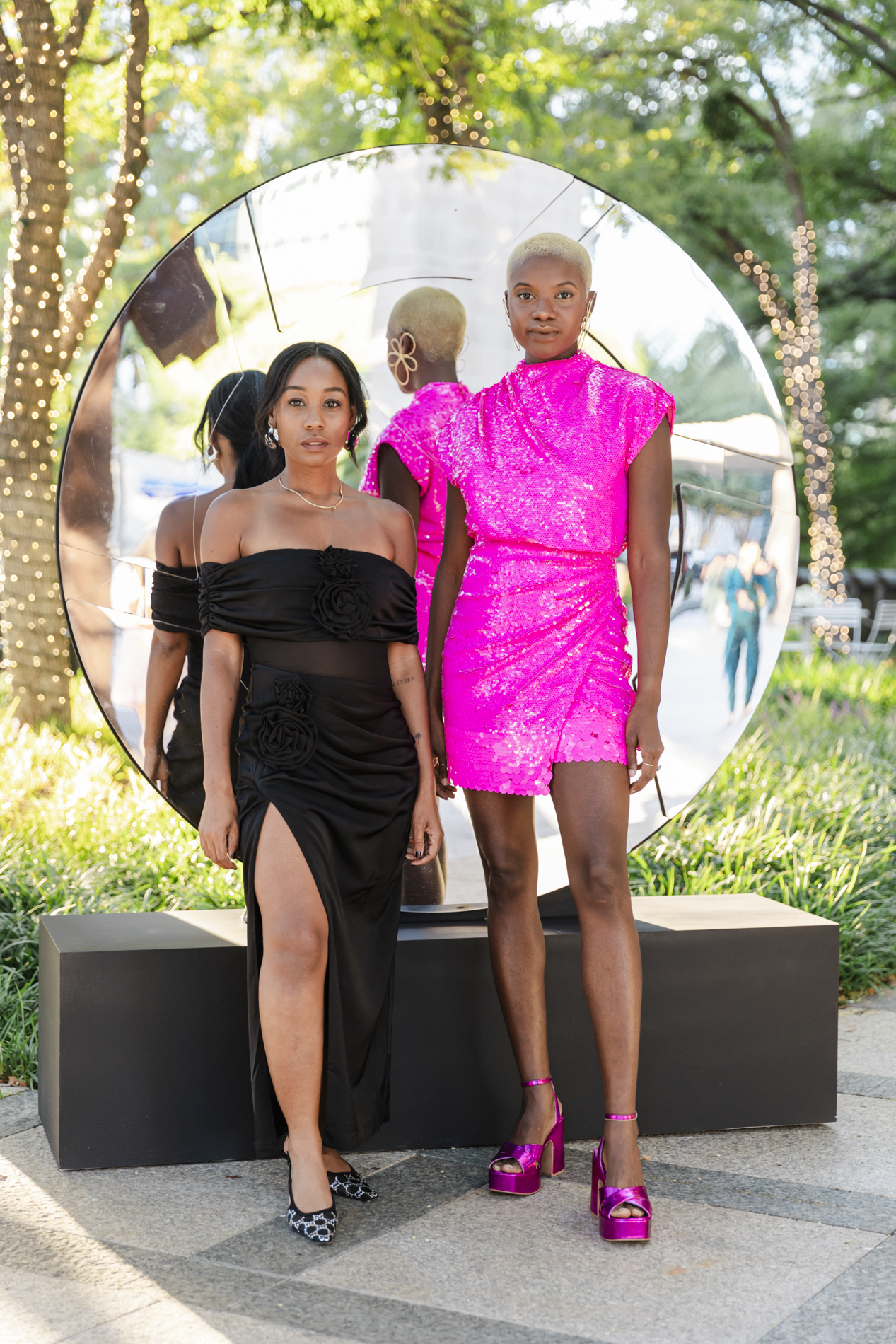 DFW event photographer: two women posing in front of a large circular mirror at an event. Both women are wearing colorful, fashionable dresses.