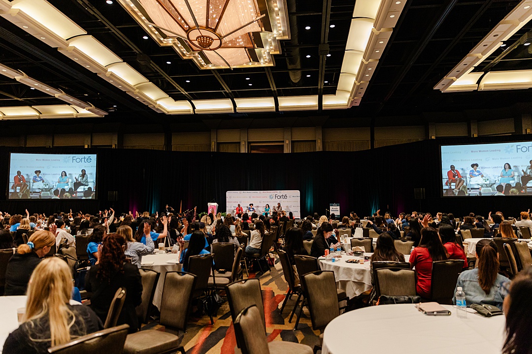 DFW Event Photographer: a wide shot of a ballroom during the Forte Foundation's MBA Women's Leadership Conference in Dallas, Texas. The foreground shows many circular ballroom tables with hundreds of women in professional business attire. The background shows a large stage with four women holding a discussion for the audience. Their image is being projected up on two large screens on either side of the stage.