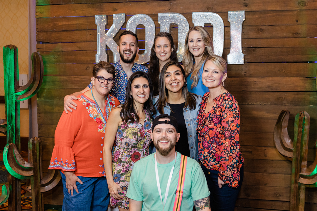 DFW Event Photography of a group of six women and two men wearing casual clothes. They are all close together and are smiling and posing in front of their company logo.