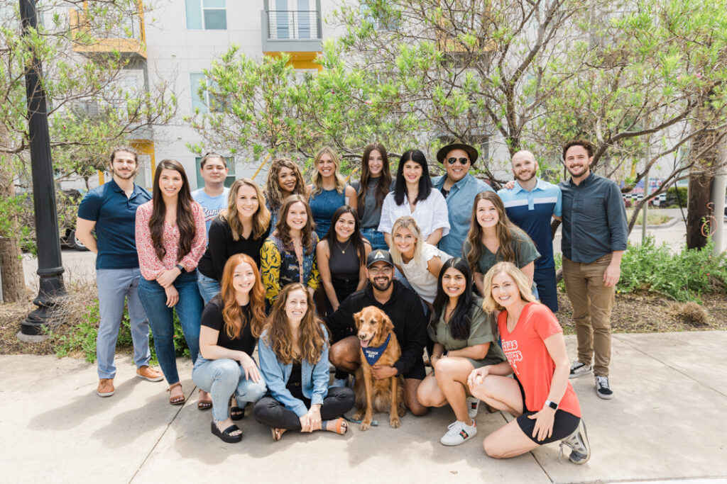 20 people wearing business casual and/or casual attire and one dog pose as a group outside on the sidewalk in front of trees and an urban setting 