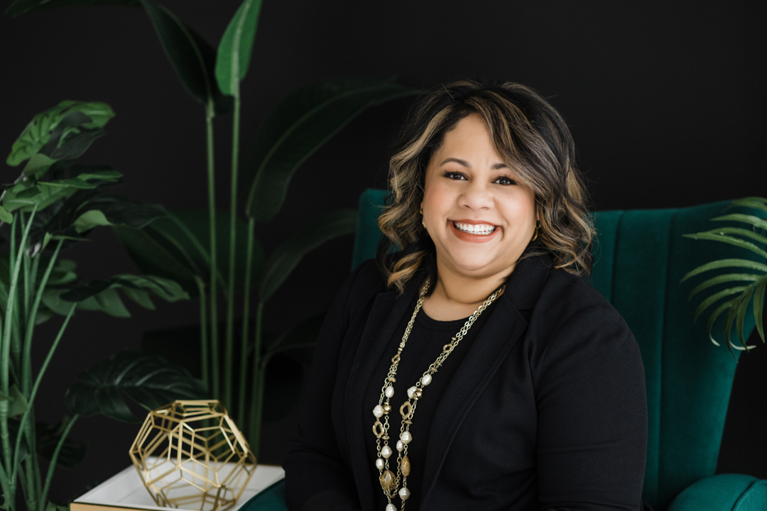 DFW Headshot Photographer; African American woman wearing all black with gold necklaces sitting in a green chair next to a gold geometric sculpture and plants in front of a black backdrop