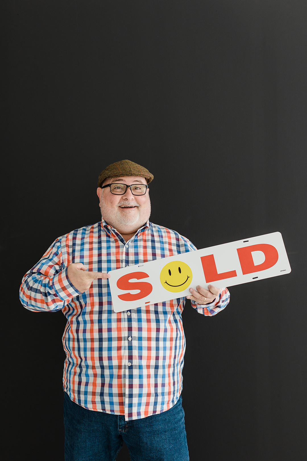 DFW Headshot Photographer; photo of a man in a blue and orange checkered shirt, brown hat, and glasses holding a sign reading "sold" and smiling in front of a black background.