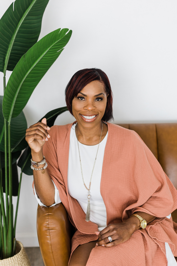 Dallas head shot photographer; African American woman wearing business casual attire, a long necklace,  bracelets, and a gold watch sitting on a leather couch next to a plant in front of a white backdrop