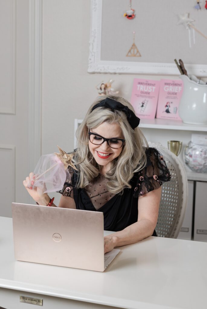 Smiling, caucasian woman wearing a black blouse, black glasses, and a black headband while working on a laptop and holding a magic wand