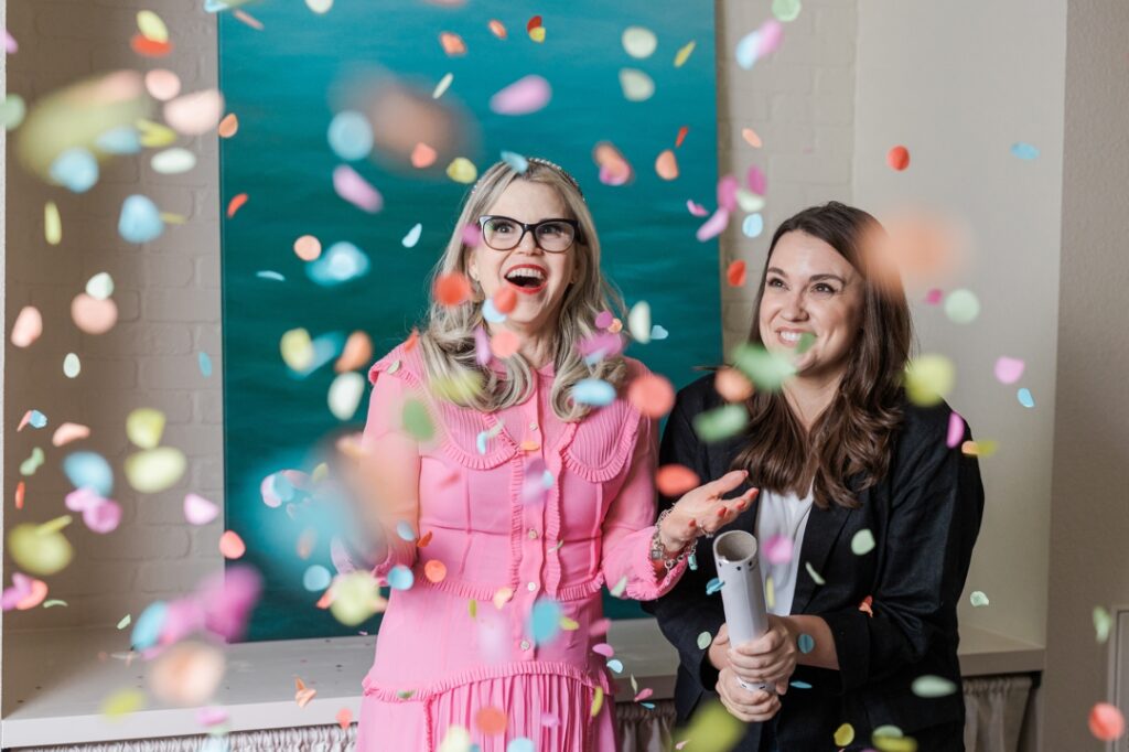 Two smiling caucasian women (one in pink and one in black and white) looking happiy at confetti they just fired out of a confetti cannon in front of a blue backdrop