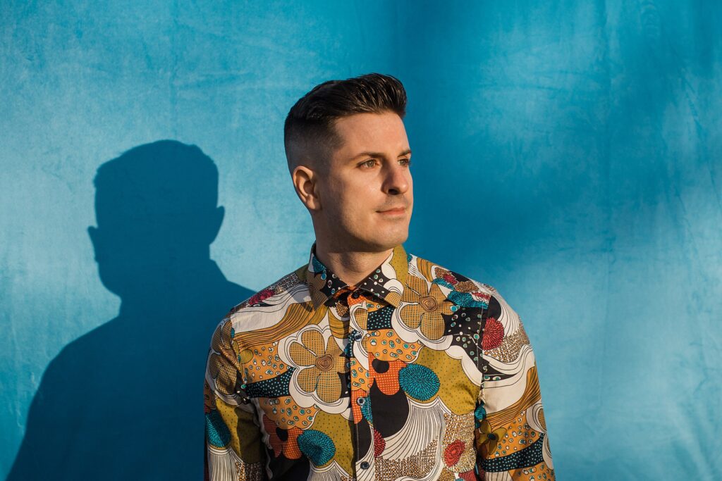 Caucasian male pensively looking off into the distance in front of a blue background. He has short dark hair and is wearing a highly detailed/colorful floral dress shirt.