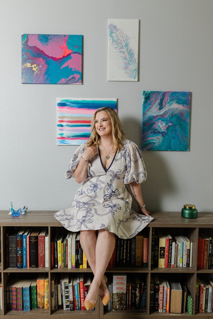 Blonde, Caucasian woman smiling and looking off to her right side. She's wearing a white dress decorated with a detailed blue pattern, a silver necklacke, and a bracelet. She is sitting in front of two abstract paintings.