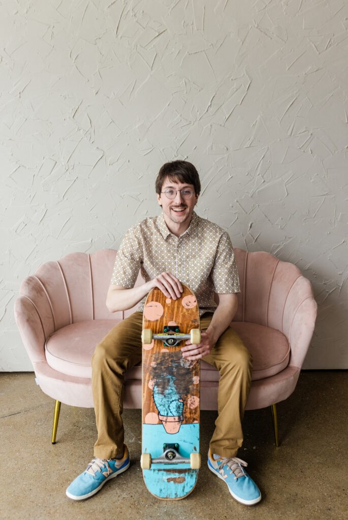 Caucasian man wearing glasses, a white and brown elaboratly patterned shirt, brown pants and blue sneakers, sitting on a salmon colored couch and holding a worn down skateboard