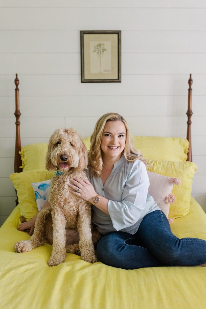 Fort Worth Brand Photographer; woman in a light blue blouse and jeans smiling and sitting with a tan dog on a yellow bed