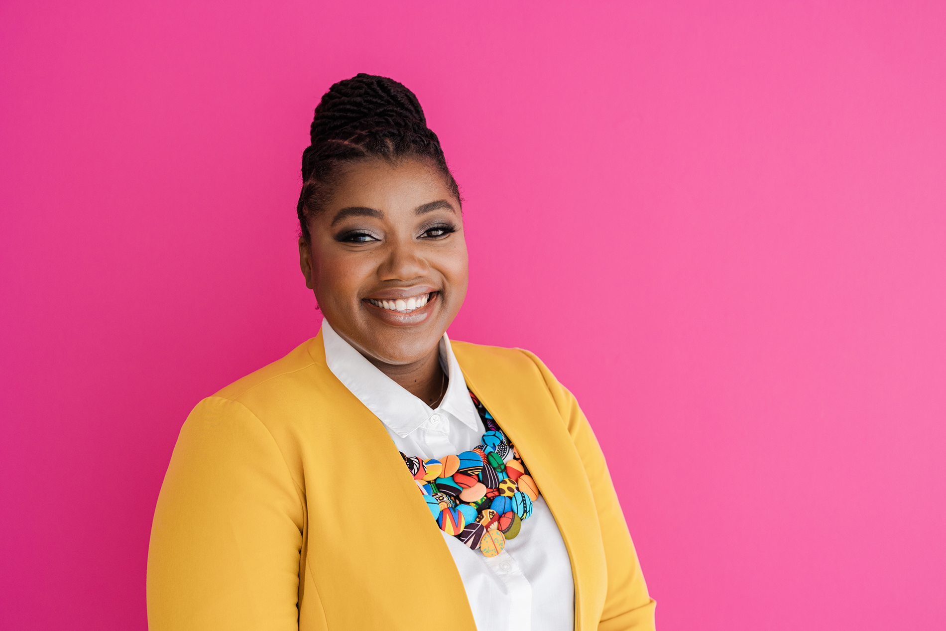 Fort Worth Branding Photographer: a woman smiling and posing in front of a pink background. She's wearing a white dress shirt, yellow blazer, and a necklace made of colorful buttons.