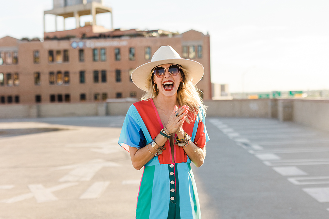 Fort Worth Branding Photographer: a woman laughing with hands together on top of a parking garage. She's wearing a multicolored jumpsuit, cowboy hat, sunglasses, and lots of jewelry.