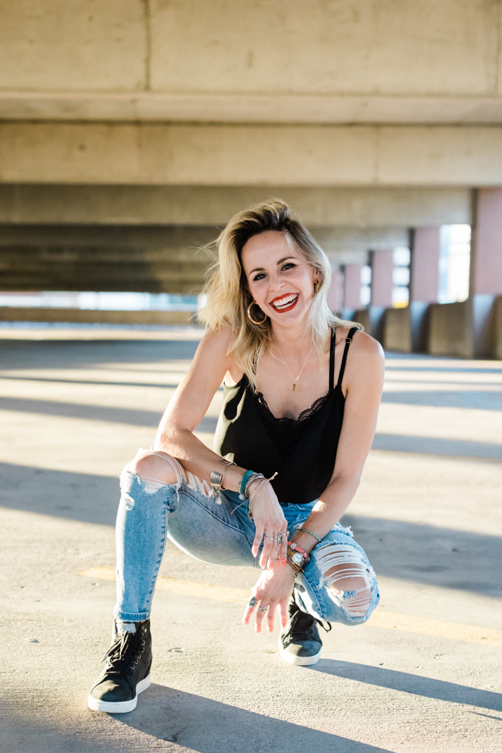 Fort Worth Editorial Brand Photographer; woman wearing a black top, ripped jeans, lots of bracelets, and sneakers smiling and kneeing in a parking garage in a downtown city setting