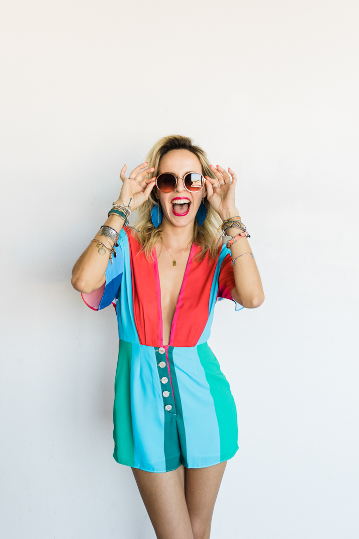 Fort Worth Editorial Brand Photographer; woman in a colorful jumpsuit, blue feather earrings, lots of bracelets, and round sunglasses smiling largely and holding her sunglasses to on her face in front of a white background