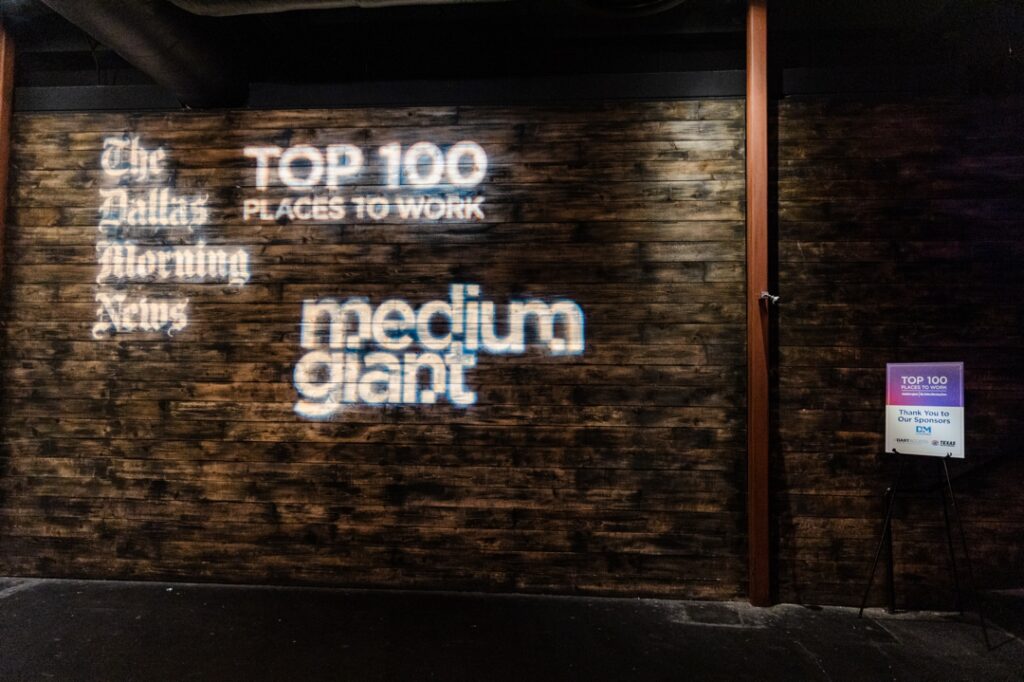 A brown, wood paneled wall with a projection of the event name, Top 100 Places to Work, and the two companies who put the event on, The Dallas Morning News and medium giant