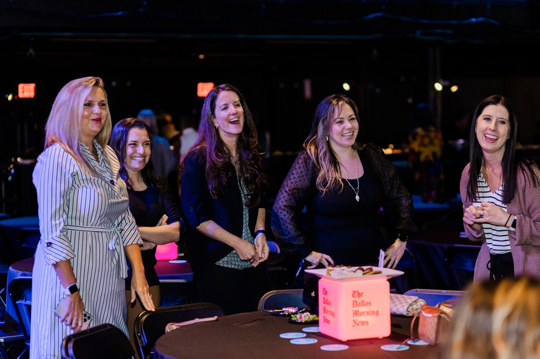 Fort Worth Event Photographer; Five women laughing and smiling and looking off to the side while standing behind an event table
