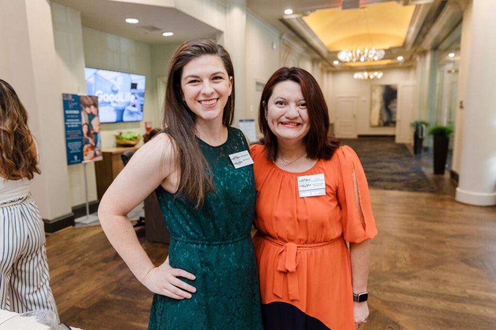 Two caucasian women members of the Society of Wedding Professionals wearing a green dress and orange dress respectively. They both smiling and posing for the camera.