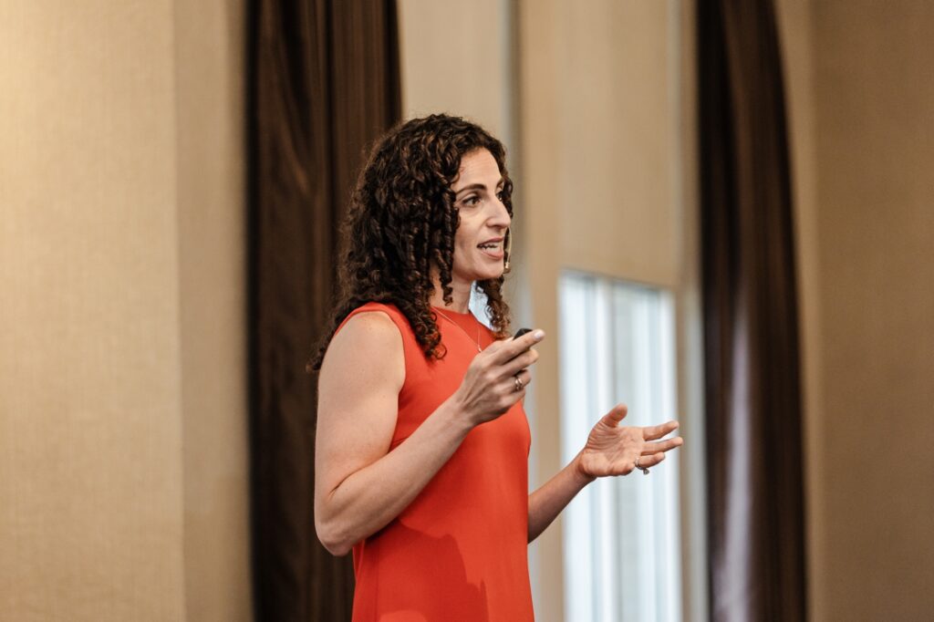 A candid shot of a member of the Society of Wedding Professionals giving a presentation while wearing an orange dress and holding a remote for the projector.