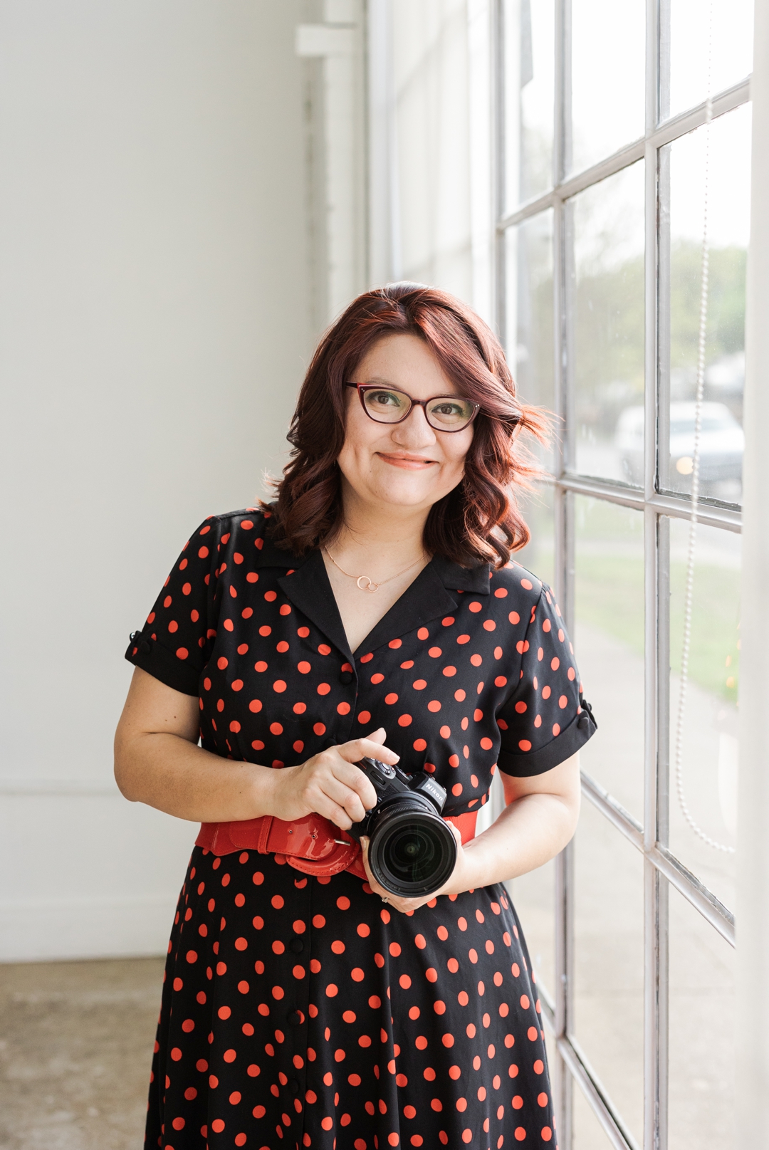 Fort Worth Headshot Photographer; Latina woman wearing a black and red polka dotted dress with a red belt and holding a Nikon DSLR camera next to a window
