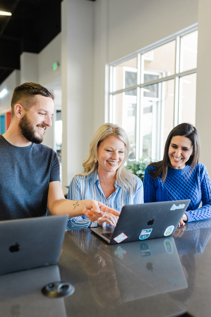 Fort Worth Office Candid Photography; candid shot of a man and two women in casual business attire smiling and looking at a laptop in an office setting