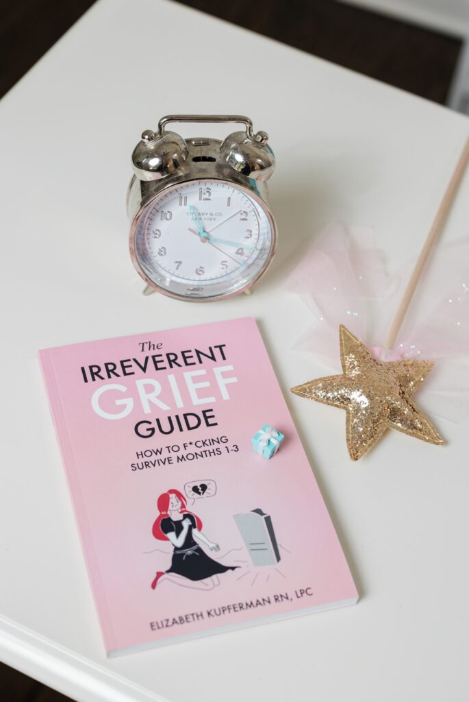 A copy of "The Irreverent Grief Guide" by Elizabeth Kupferman, a magic wand, and a traditional alarm clock all resting on a white table
