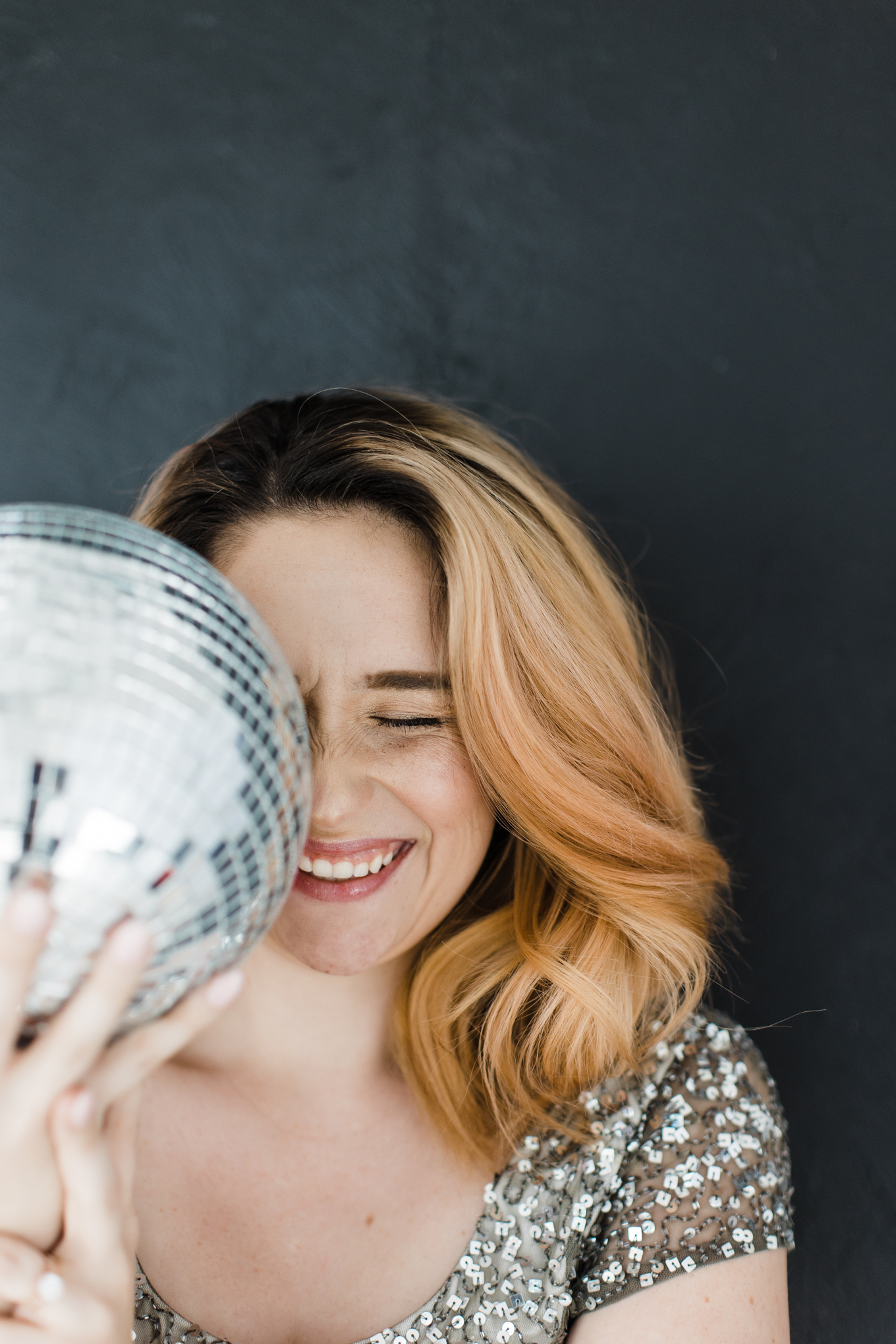 Fun Brand Photographer; photo of a woman in a sparkly, sequin, silver top smiling with closed eyes and hiding the right side of her face behind a mirrored disco ball in front of a dark background