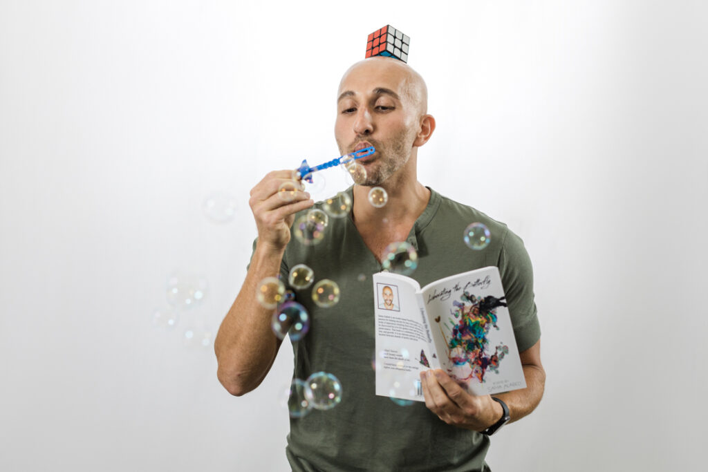 Caucasian male with a shaved head and a green shirt wearing a Rubik's cube like a hat; he is blowing bubbles with a blue bubble wand in his right hand and is holding an open book in his left hand in front of a white backdrop