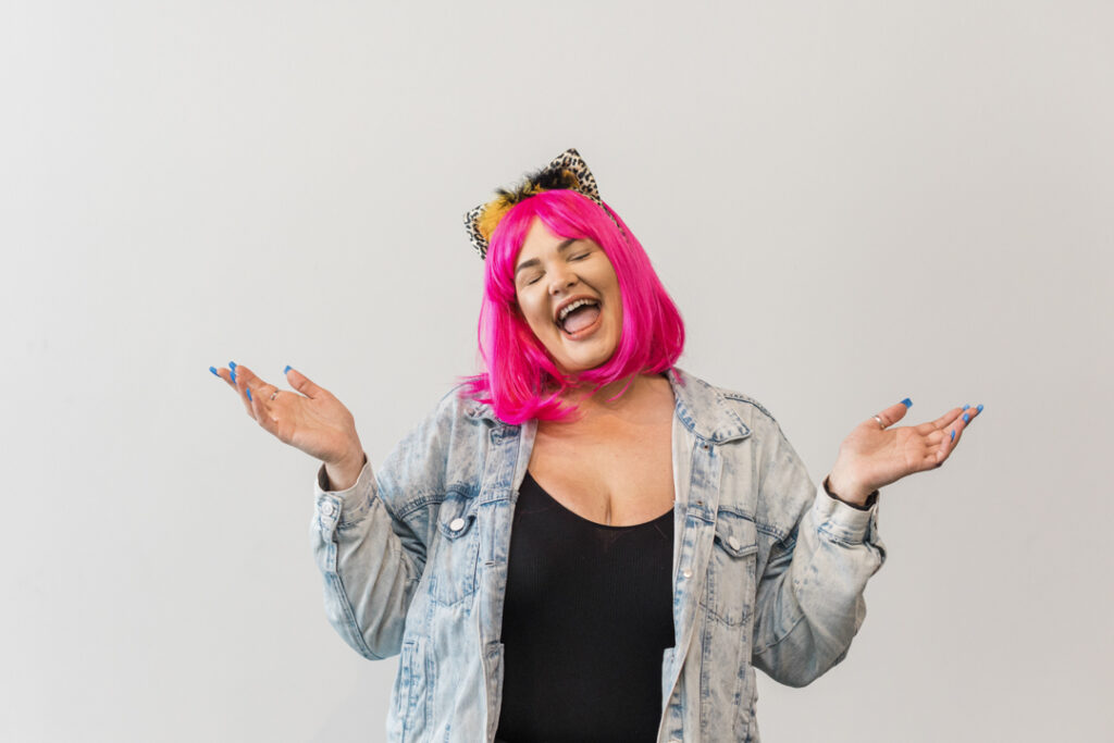 Woman with pink hair wearing a leopard print bow, a black shirt, and a denim jacket in the midst of a candid expression in front of a white backdrop