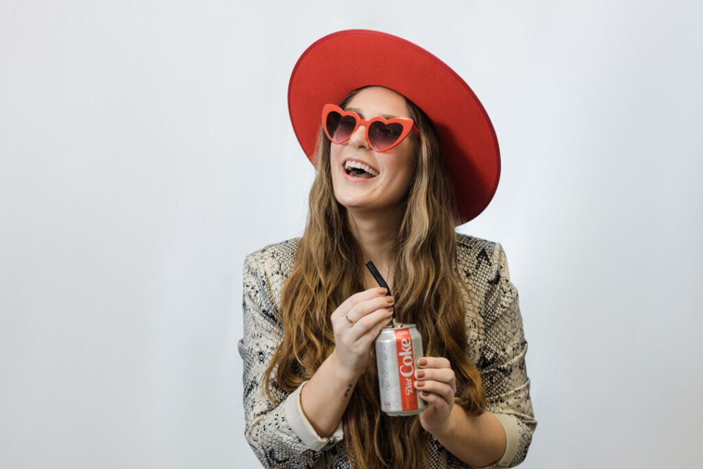 Caucasian woman with long light brown hair wearing red heart shaped sunglasses, a red hat, and a patterned jacket while holding a diet soda with a straw in front of a white backdrop
