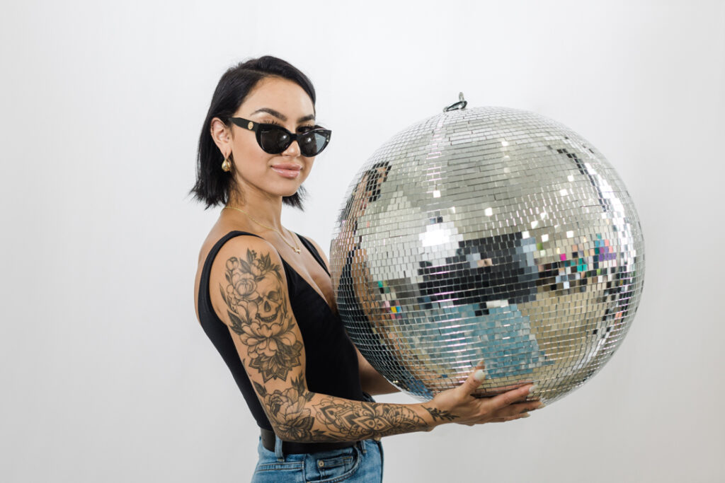 Woman with medium black hair wearing fashionable sunglasses, a black top, and jeans with a full sleeve of tattoos on her right arm; she is holding a large disco ball in front of a white backdrop