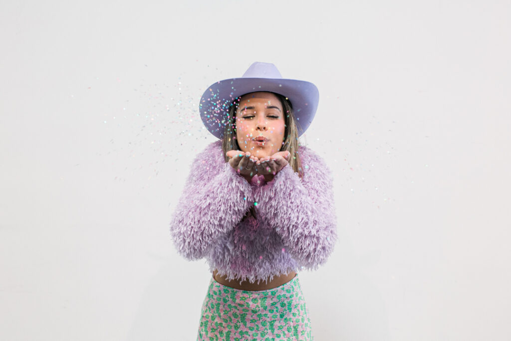 Caucasian woman with long blonde hair wearing a shag pink top, a green and pink skirt, and a lavender cowboy hat while blowing glitter towards the camera in front of a white backdrop