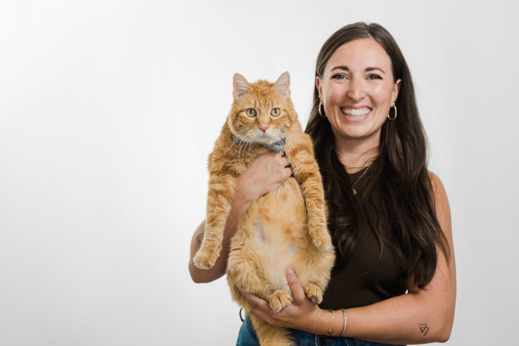Caucasian woman with long brown hair, a brown top, and jeans smiles and poses with a large orange tabby cat in front of a white backdrop