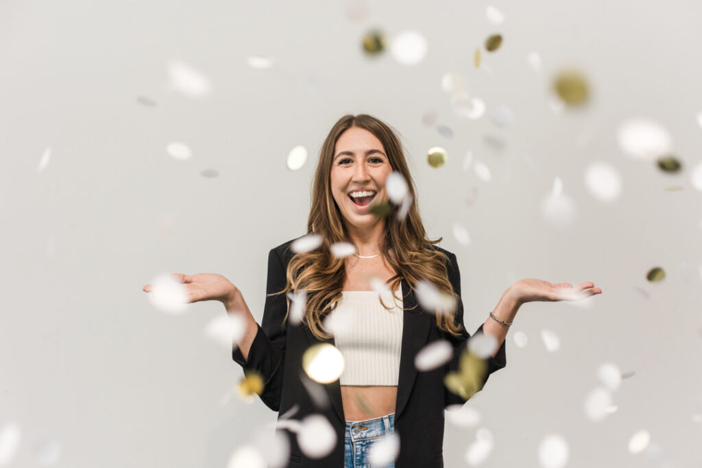 Caucasian woman with long dark blonde hair with a white top, a black jacket, and jeans throwing golden confetti into the air while smiling in front of a white backdrop