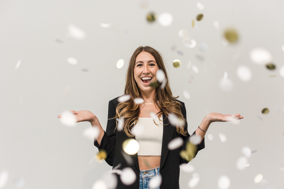 Fun DFW Headshot Photographer; photo of a woman in jeans, jacket, and crop top smiling and throwing silver and gold confetti in front of a white background