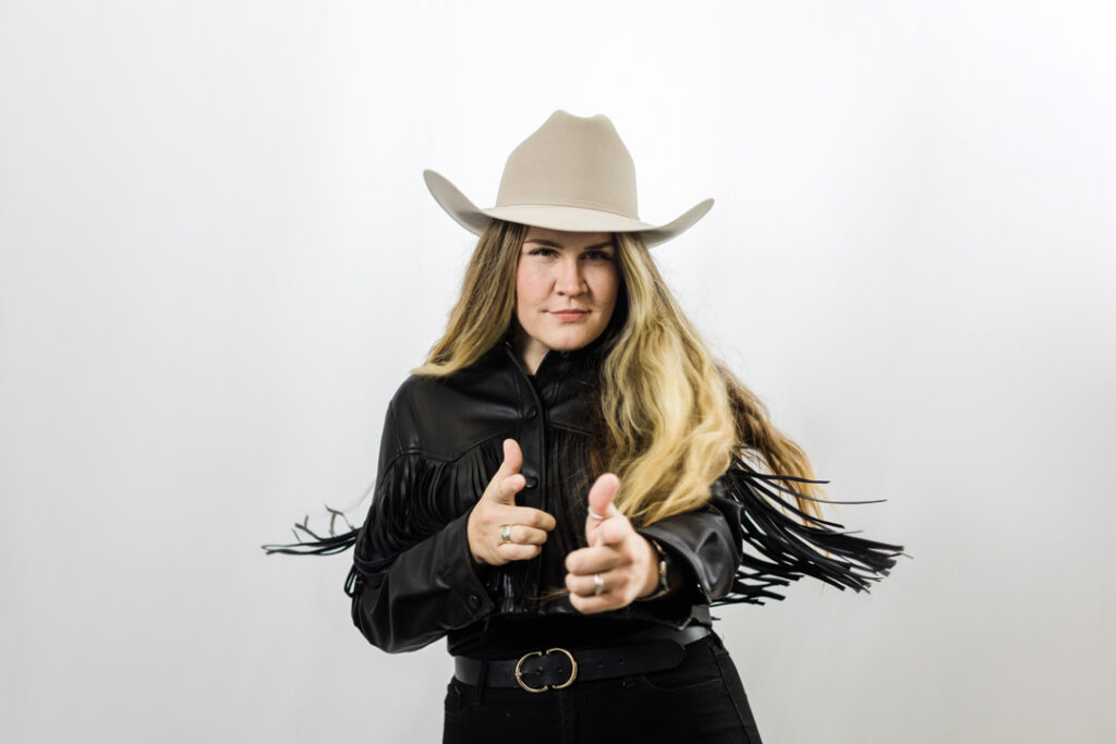 Caucasian woman with long blonde hair wearing black western attire and a white cowboy hat posing with finger guns in front of a white backdrop