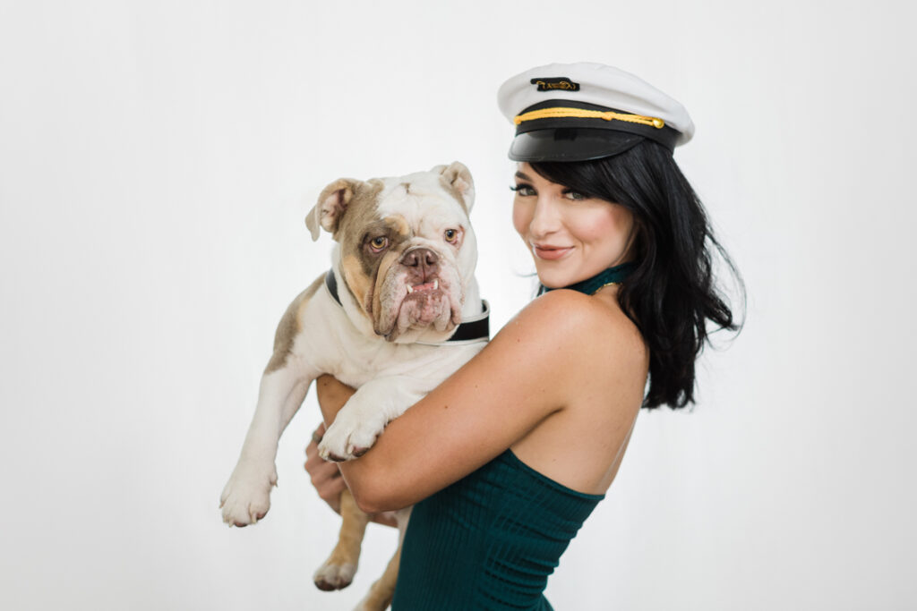 Caucasian woman with long black hair wearing a captain's hat and a green shirt holding a brown and white dog in front of a white backdrop