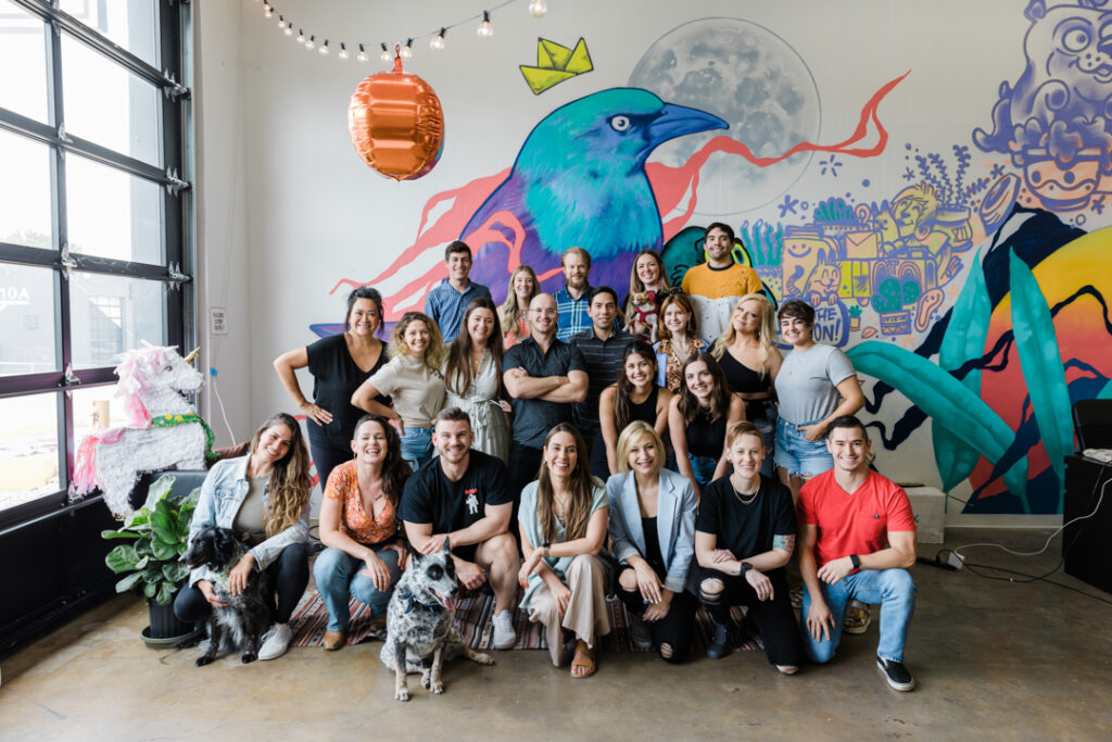 22 people and 2 dogs posing as a large group in front of a colorful mural next to a large window with a unicorn pinata
