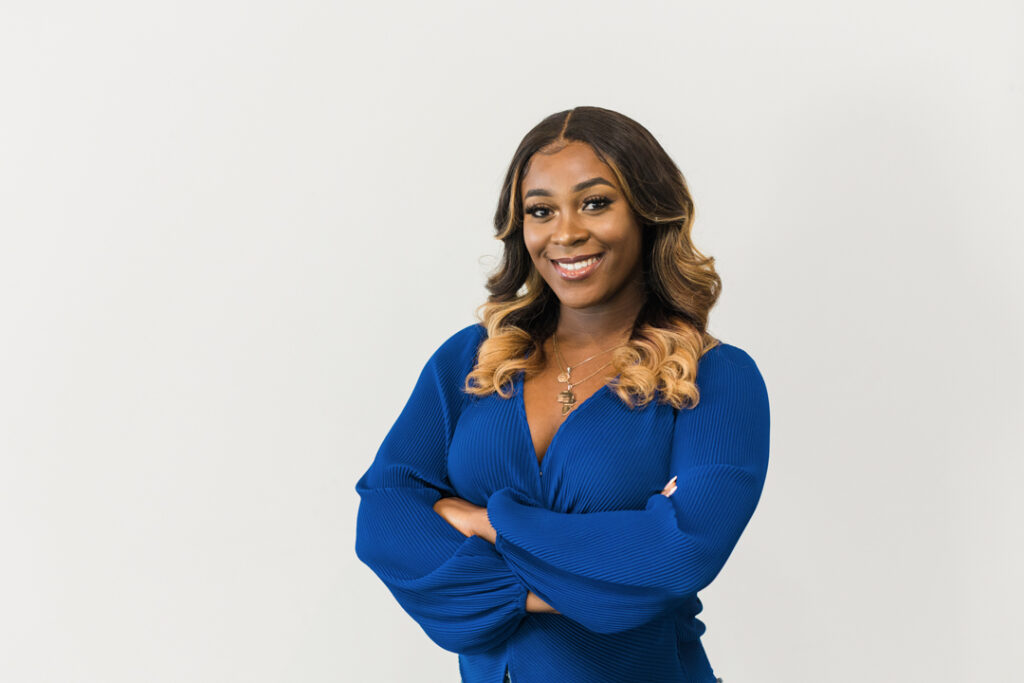 African American woman with long brown hair with highlights wearing a blue dress with multiple necklaces posing with her arms crossed in front of a white backdrop