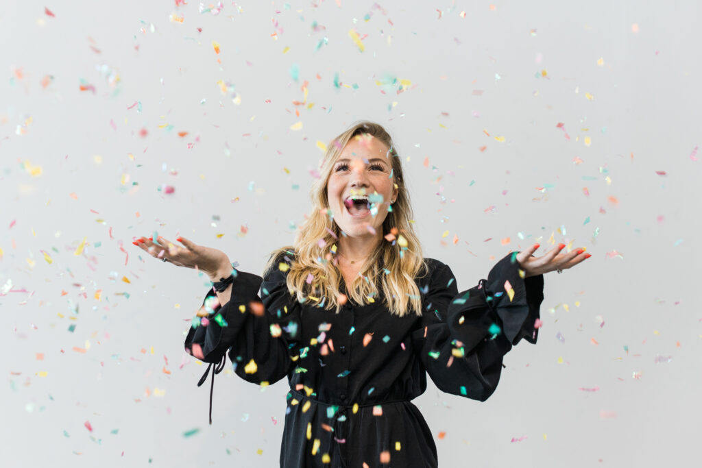 Caucasian woman with blonde hair in a black dress looking joyfully at colorful confetti she tossed into the air in front of a white backdrop