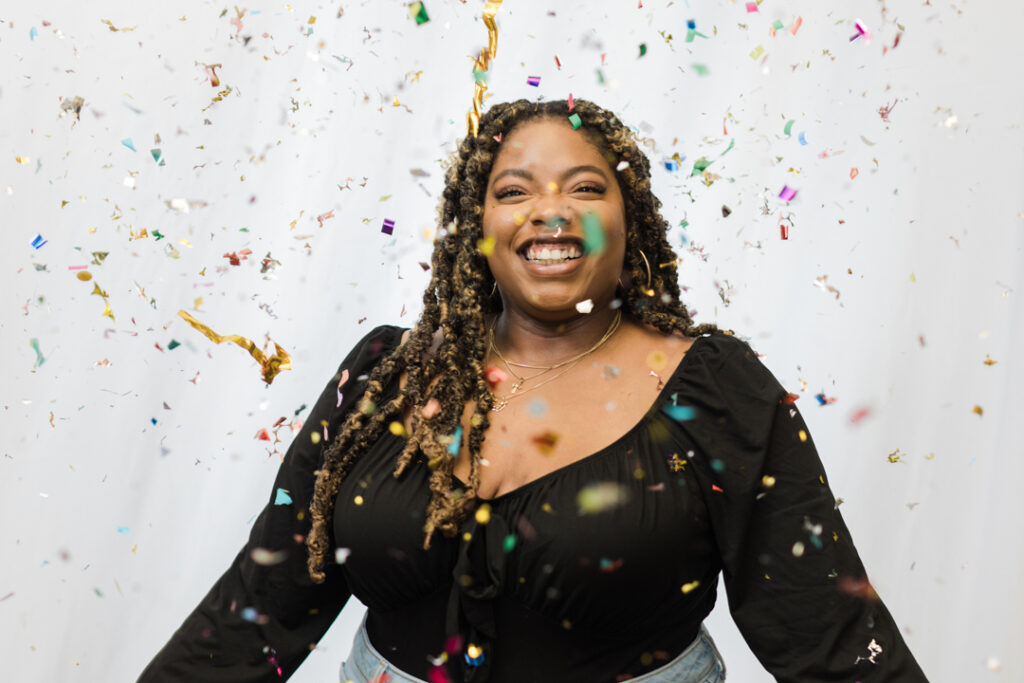 African American woman with long curly brown hair wearing a black top and jeans smiling while confetti falls around her in front of a white backdrop